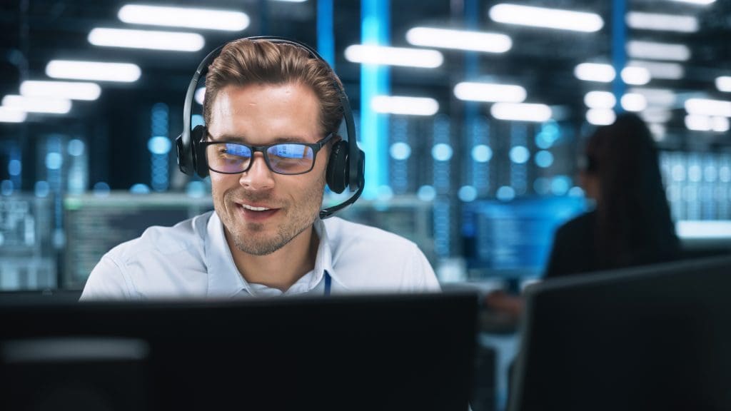 A man wearing a headset and glasses and helping someone remotely on a computer.